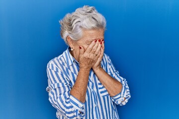 Senior woman with grey hair standing over blue background with sad expression covering face with hands while crying. depression concept.