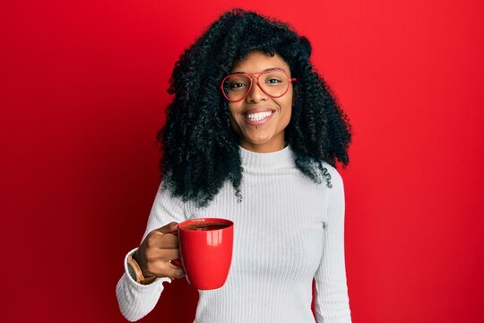 African american woman with afro hair holding coffee looking positive and happy standing and smiling with a confident smile showing teeth