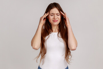 Woman with dark hair frowning, clasping sore head, suffering intense headache, having unbearable migraine, fever and flu symptoms, wearing white T-shirt. Indoor studio shot isolated on gray background