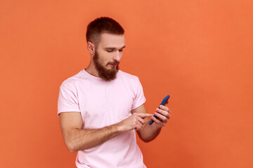 Portrait of bearded man scrolling online, texting message in social media on cell phone, using mobile network services, wearing pink T-shirt. Indoor studio shot isolated on orange background.