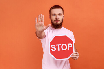 Portrait of strict bossy bearded man showing stop gesture and holding red stop road sign, prohibitions and restrictions, wearing pink T-shirt. Indoor studio shot isolated on orange background.