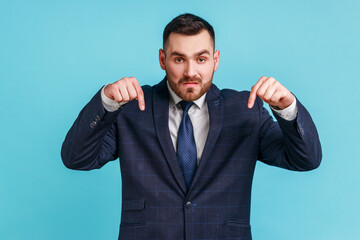 Come to me here now! Young adult bearded man in suit pointing fingers down, demanding approach to him immediately, having control over situation. Indoor studio shot isolated on blue background.