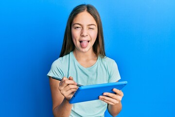 Beautiful brunette little girl using touchpad drawing on screen sticking tongue out happy with...