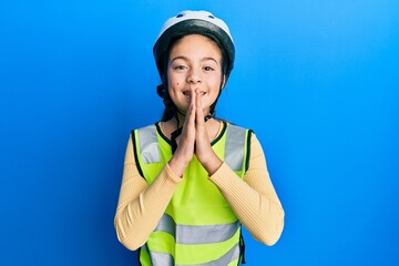 Beautiful brunette little girl wearing bike helmet and reflective vest praying with hands together asking for forgiveness smiling confident.