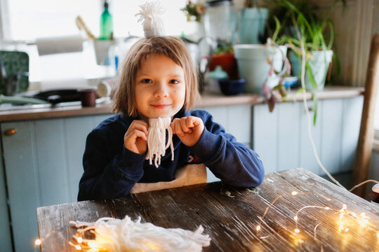 A cute caucasian child makes a craft from threads, a girl at a wooden table makes hats from yarn, a child and creativity at home. Dark style, lifestyle in a real interior.