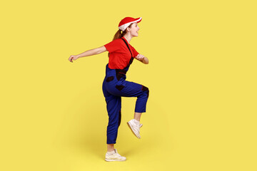 Fototapeta na wymiar Side view portrait of worker woman marching, going for work, expressing positive emotions and happiness, wearing overalls and red cap. Indoor studio shot isolated on yellow background.