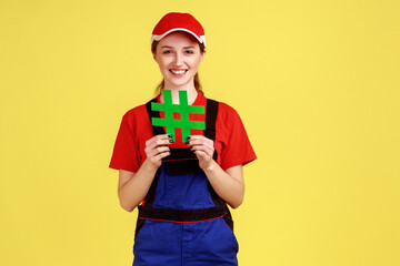 Portrait of smiling handy woman standing holding green hashtag in hands, looking at camera with happy expression, wearing overalls and red cap. Indoor studio shot isolated on yellow background.
