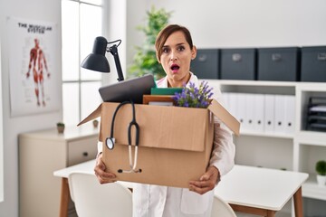 Middle age doctor woman holding cardboard box with items at the clinic afraid and shocked with surprise and amazed expression, fear and excited face.