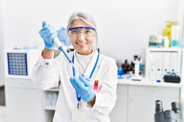 Middle age grey-haired woman wearing scientist uniform using pipette at laboratory