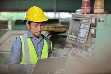 Technician engineer or worker in protective uniform standing and using computer while controlling operation or checking industry machine process with hardhat  at heavy industry manufacturing factory