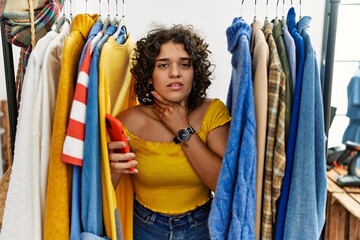 Fototapeta na wymiar Young hispanic woman searching clothes on clothing rack using smartphone touching painful neck, sore throat for flu, clod and infection