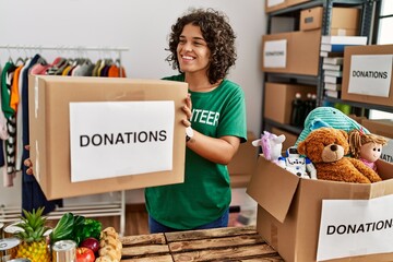 Young hispanic woman wearing volunteer uniform holding donations box at charity center