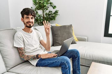 Hispanic man with beard sitting on the sofa showing and pointing up with fingers number four while smiling confident and happy.