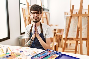 Hispanic man with beard at art studio begging and praying with hands together with hope expression...