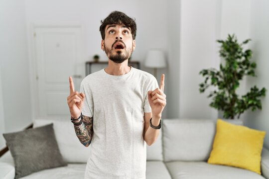 Hispanic man with beard at the living room at home amazed and surprised looking up and pointing with fingers and raised arms.