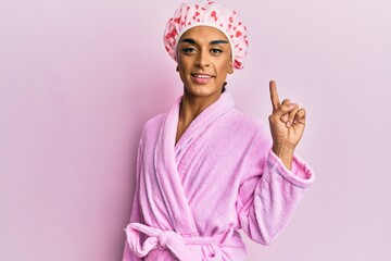 Hispanic man wearing make up wearing shower towel cap and bathrobe showing and pointing up with finger number one while smiling confident and happy.