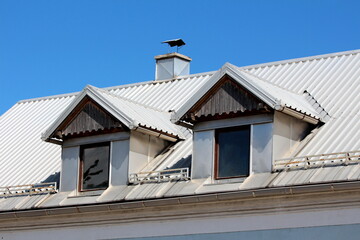 Two old roof windows with wooden frames and wooden dilapidated decorative boards on top covered with newly installed metal roof tiles surrounded with snow guards and metal sheets covered chimney