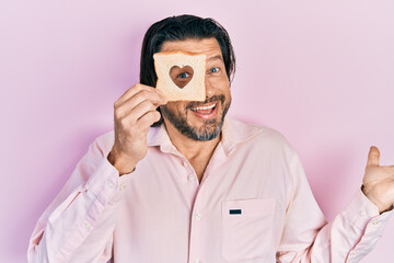 Middle age caucasian man holding bread loaf with heart shape celebrating achievement with happy smile and winner expression with raised hand