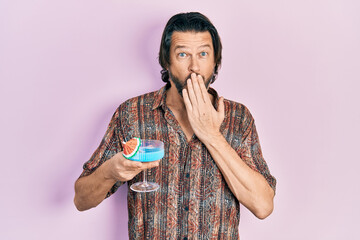 Middle age caucasian man drinking tropical cocktail with alcohol covering mouth with hand, shocked and afraid for mistake. surprised expression