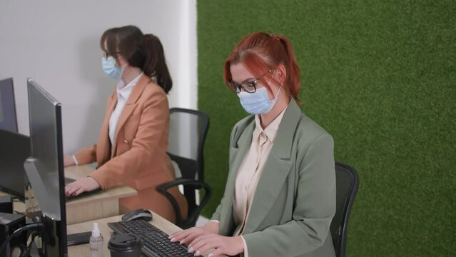 workplace precautions, young female employees in medical masks comply with quarantine measures and use an antiseptic to treat surfaces and hands from a dangerous virus while sitting at a table indoor