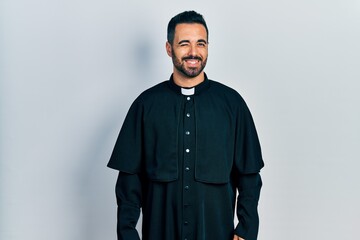 Handsome hispanic man with beard wearing catholic priest robe winking looking at the camera with...