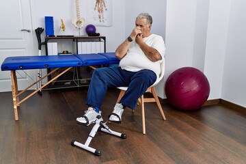 Fototapeta na wymiar Senior caucasian man at physiotherapy clinic using pedal exerciser smelling something stinky and disgusting, intolerable smell, holding breath with fingers on nose. bad smell