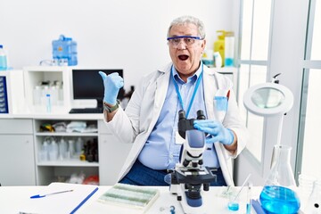 Senior caucasian man working at scientist laboratory surprised pointing with hand finger to the side, open mouth amazed expression.