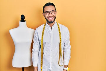 Hispanic man with beard dressmaker designer standing by manikin looking positive and happy standing and smiling with a confident smile showing teeth