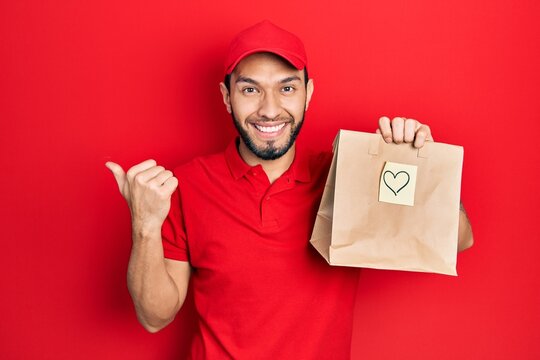 Hispanic man with beard holding take away paper bag with heart reminder pointing thumb up to the side smiling happy with open mouth