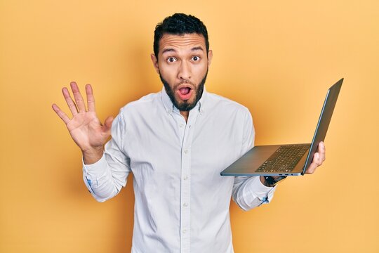 Hispanic man with beard working using computer laptop scared and amazed with open mouth for surprise, disbelief face