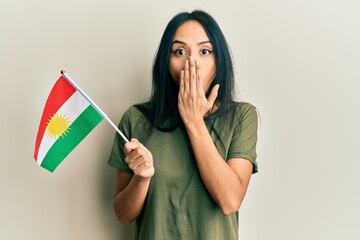 Young hispanic girl holding kurdistan flag covering mouth with hand, shocked and afraid for...