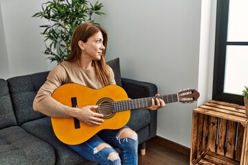 Middle age caucasian woman playing classical guitar sitting on the sofa at home.