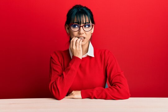 Young brunette woman with bangs wearing glasses sitting on the table looking stressed and nervous with hands on mouth biting nails. anxiety problem.