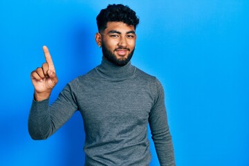 Fototapeta premium Arab man with beard wearing turtleneck sweater pointing finger up with successful idea. exited and happy. number one.