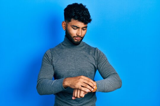 Arab man with beard wearing turtleneck sweater checking the time on wrist watch, relaxed and confident