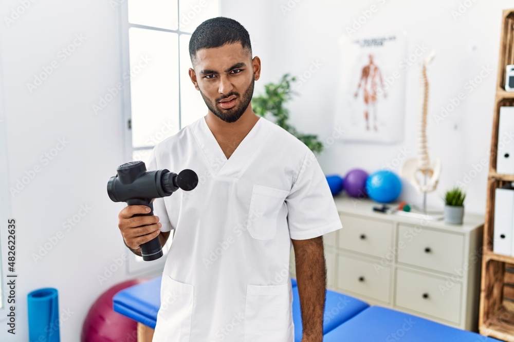 Poster Young indian physiotherapist holding therapy massage gun at wellness center in shock face, looking skeptical and sarcastic, surprised with open mouth