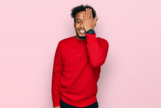 Young african american man with beard wearing casual winter sweater covering one eye with hand, confident smile on face and surprise emotion.