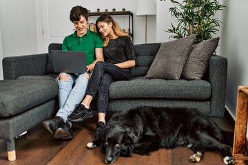 Young caucasian couple smiling happy using laptop sitting on the sofa with dog at home.