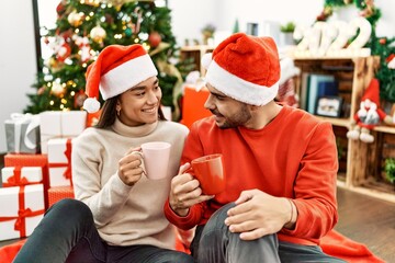 Fototapeta premium Young hispanic couple smiling happy and drinking cup of coffee sitting on the floor by christmas tree at home.