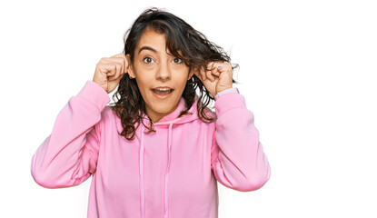Young hispanic woman wearing casual sweatshirt smiling pulling ears with fingers, funny gesture....