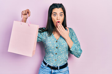 Young hispanic girl holding shopping bags covering mouth with hand, shocked and afraid for mistake....