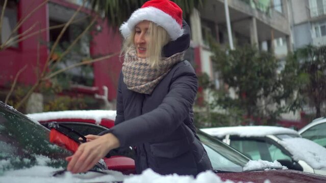 A young beautiful Caucasian woman in a Santa Claus hat cleans a car from snow with a brush in the yard of her house against a background of palm trees and trees. She is joyful and happy with a smile.