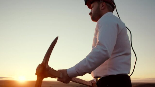 Man stands background sunset in helmet and throws pickaxe over shoulder, side view. Businessman in hard hat stands outdoor and throws pickaxe over shoulder