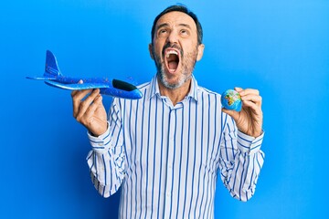 Middle age hispanic man holding plane toy and world ball angry and mad screaming frustrated and furious, shouting with anger looking up.