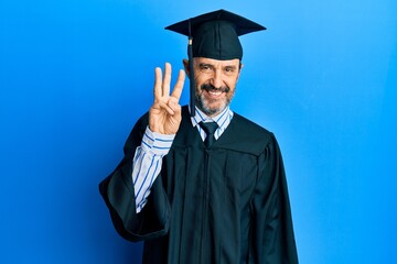 Middle age hispanic man wearing graduation cap and ceremony robe showing and pointing up with fingers number three while smiling confident and happy.