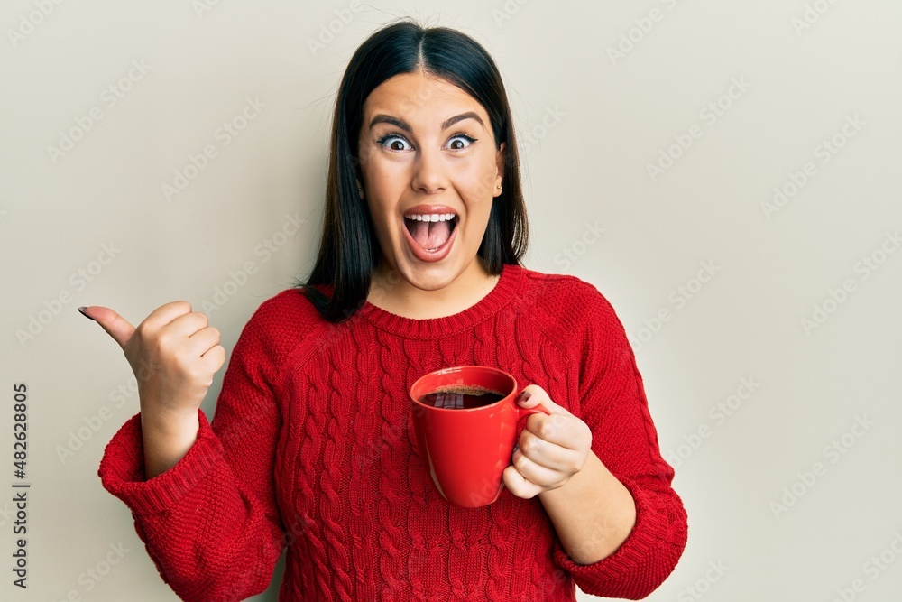 Poster beautiful brunette woman drinking a cup of black coffee pointing thumb up to the side smiling happy 