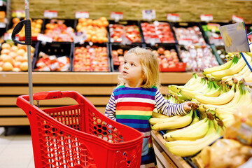 Cute todler girl pushing shopping cart in supermarket. Little child buying fruits. Kid grocery shopping. Adorable baby kid with trolley choosing fresh vegetables in local store.