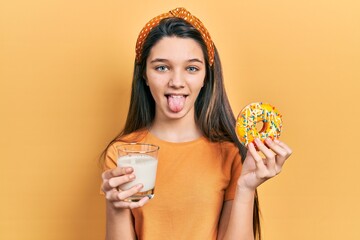 Young brunette girl eating donut and drinking glass of milk sticking tongue out happy with funny...