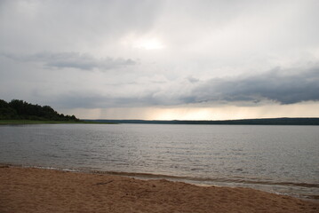 Wide flood of the Vuoksa river. Beach brown sand, river with clear clean water on it small waves. From above, white-gray with blue clouds from which it is raining in the distance.