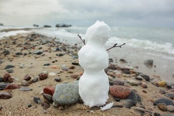 tiny snowman with branches hands raised up stands on a sandy beach among the pebbles and looks at...